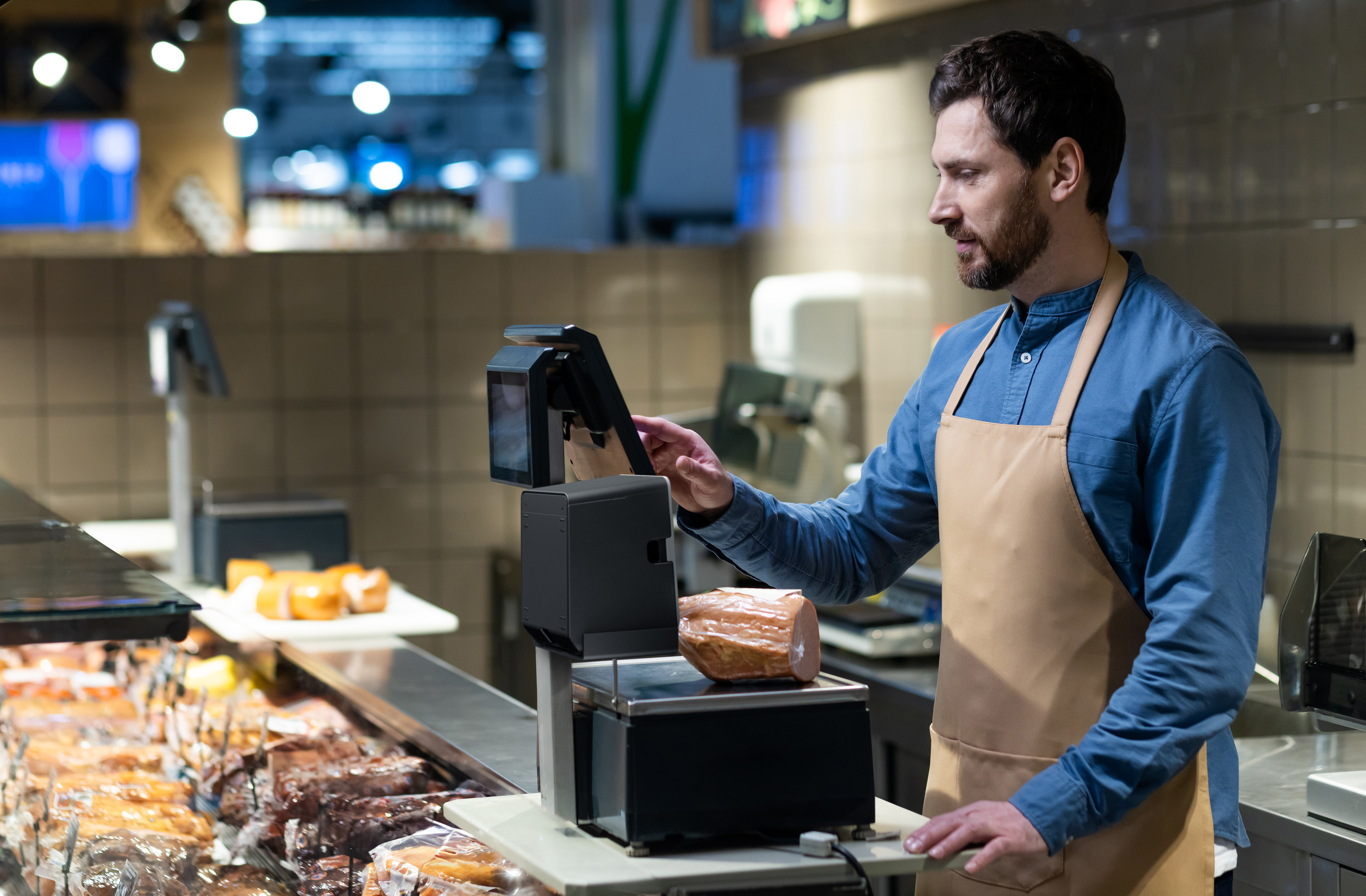 Butcher in grocery store weighing deli meats while preparing customer orders behind counter with digital scales and packaged products